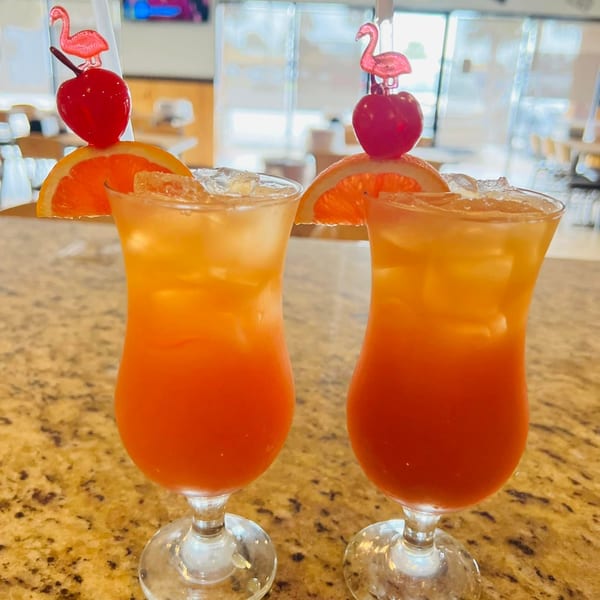 Two vibrant orange and red cocktails with orange slices, cherries, and flamingo stirrers on a bar.